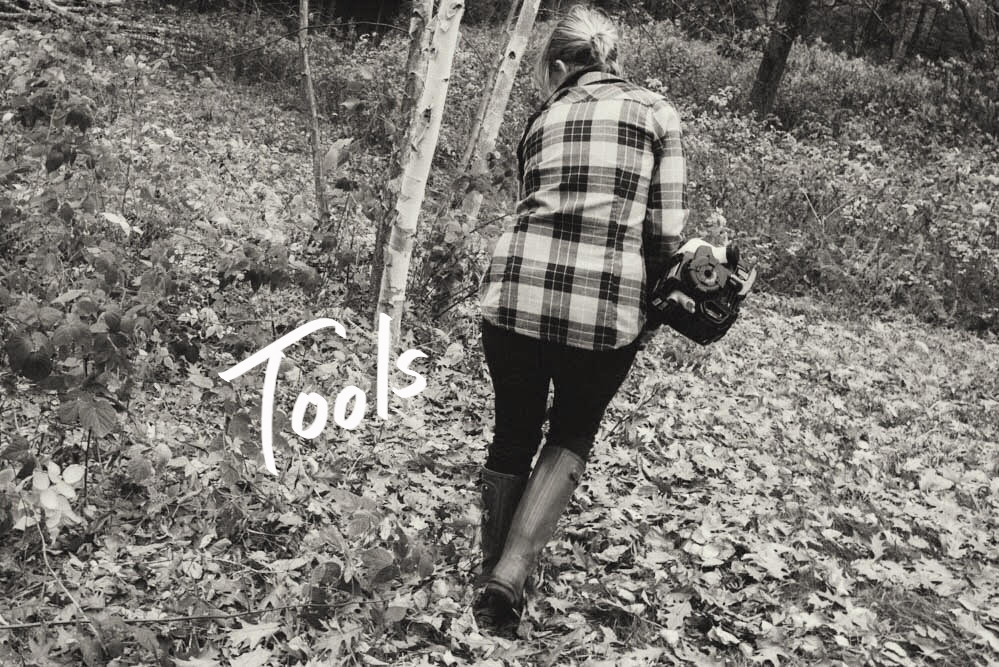 A black and white photo of a woman walking in the woods, carrying tools.