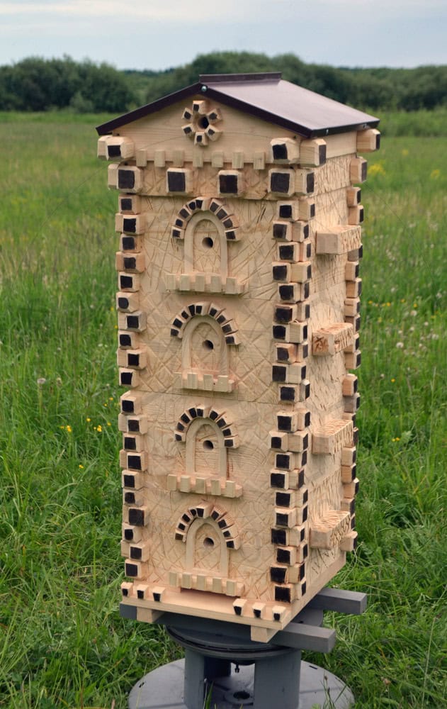 A tall, intricately carved wooden structure resembling a medieval tower stands in the middle of a grassy field. This bee hive art piece features detailed arches and decorative elements, with small ledges protruding from its sides. A metal base supports the structure.