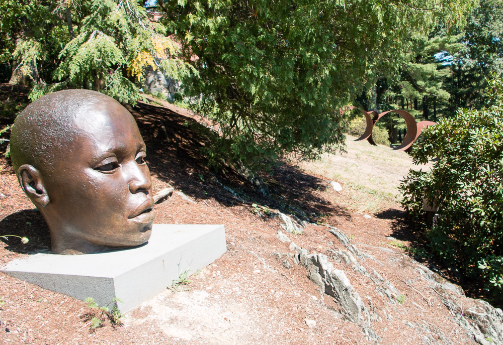 A large bronze head sculpture of a contemplative person rests on a concrete pedestal outdoors, surrounded by trees and vegetation at the deCordova Museum. In the background, a rust-colored metal circular sculpture is partially visible in a grassy field.