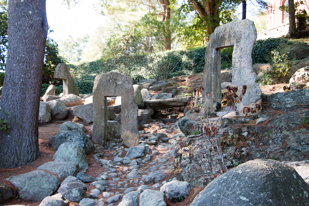 A tranquil forest scene resembling the Decordova Museum grounds features ancient stone structures with rectangular openings, surrounded by rocks, trees, and light vegetation. Sunlight filters through the trees, casting dappled shadows on the ground and stones.