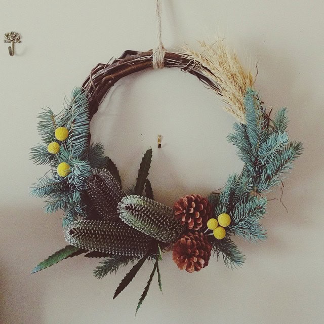 A decorative holiday wreath with blue spruce branches, yellow billy balls, pinecones, and green foliage hangs elegantly on the wall. The wreath showcases a natural, rustic design with a delightful mix of textures and colors.