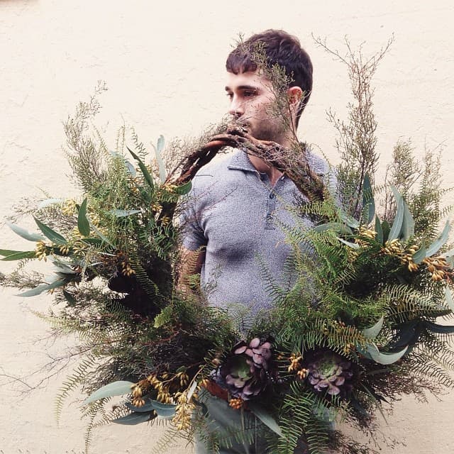 A person in a gray shirt stands against a light wall, holding a large, lush holiday wreath adorned with various green foliage and succulents. The wreath partially obscures the person's face, adding an artistic, natural feel to the image.