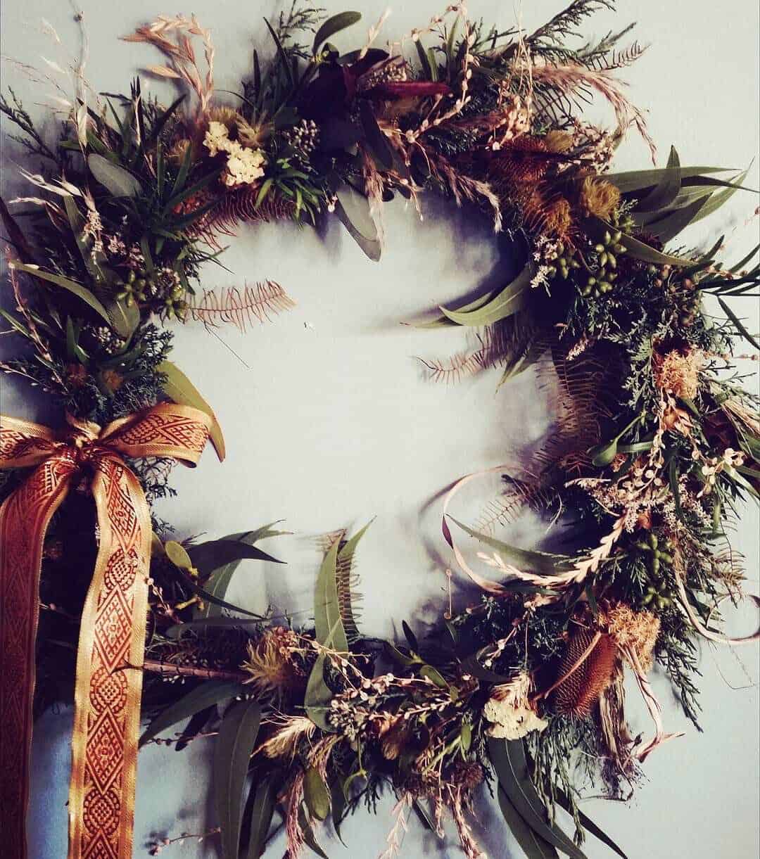 A decorative holiday wreath made of various green and brown foliage, accented with small flowers, hangs against a light-colored wall. A patterned ribbon is tied into a bow on the bottom left of the wreath.