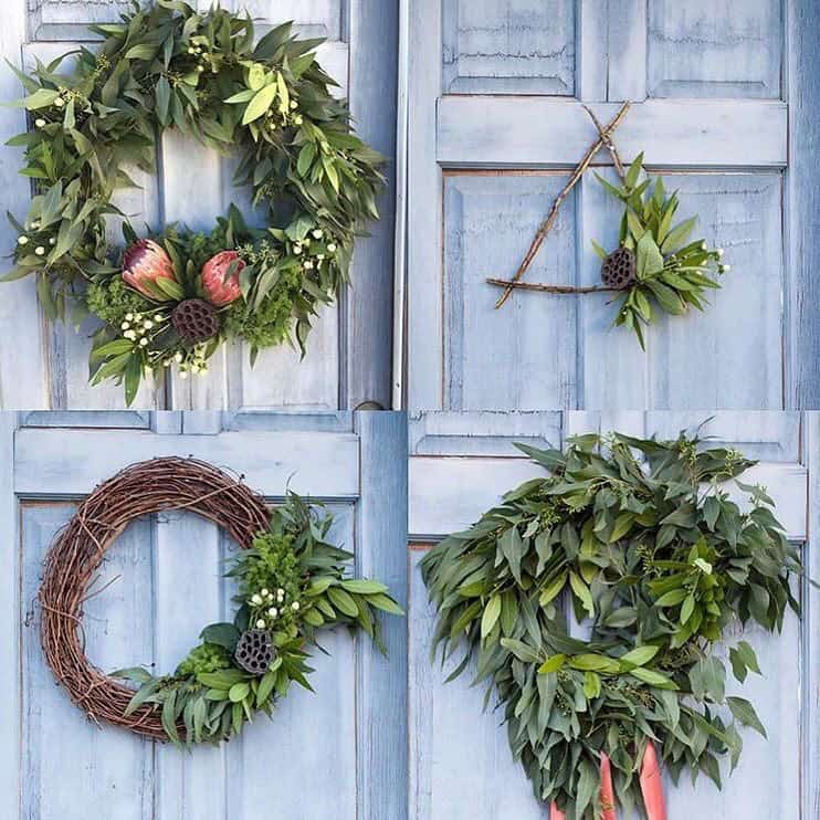 Four wreaths hang on a blue door: Top left features protea and eucalyptus, perfect for a holiday wreath. Top right is minimalist with twigs. Bottom left is round with greenery, while the bottom right is lush with eucalyptus and adorned with festive red ribbons.