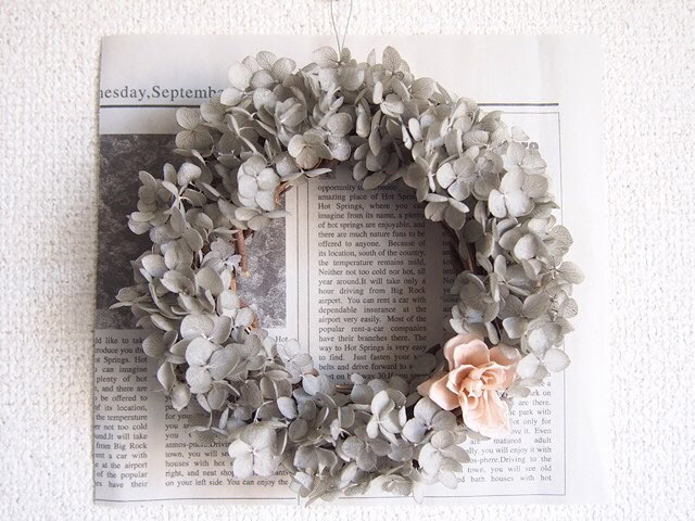 A holiday wreath crafted from pale, dried leaves and adorned with a single light pink flower hangs elegantly on the wall. Placed over a newspaper page, it exudes a charming rustic or vintage decorative theme.