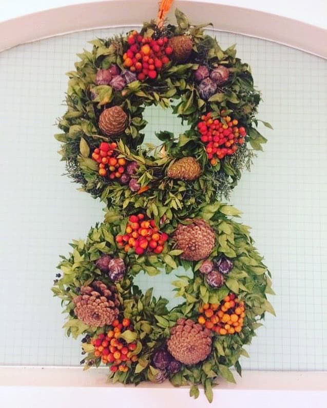 Two holiday wreaths are stacked vertically, adorned with green leaves, pine cones, orange berries, and small purple cabbages. They hang gracefully against a grid-patterned background.