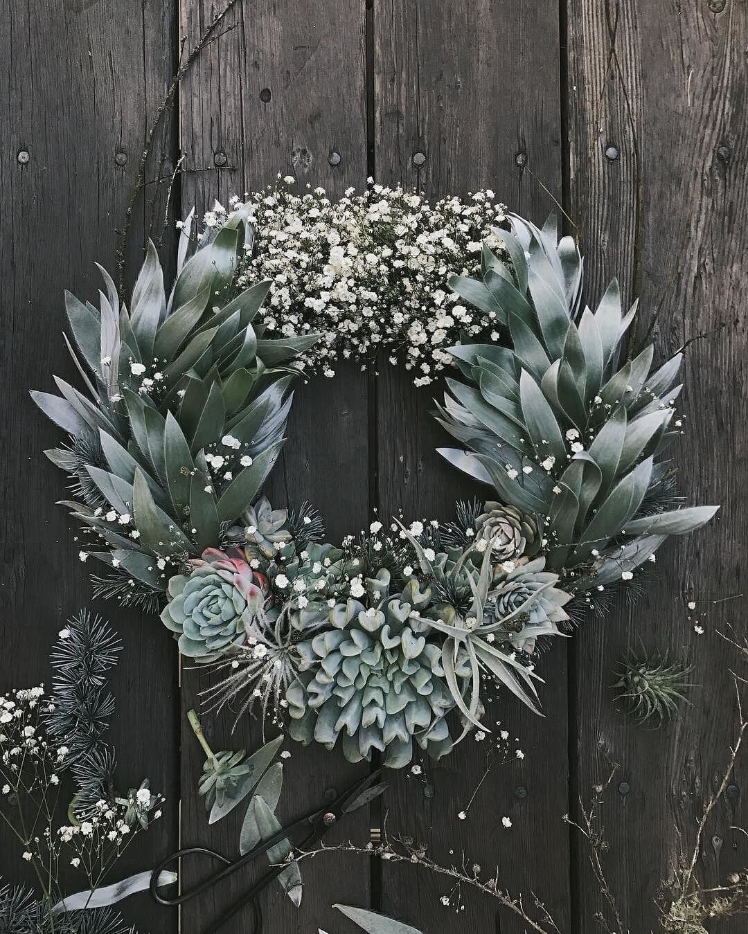 A circular holiday wreath on a dark wooden background, adorned with green succulents, eucalyptus leaves, and small white baby's breath flowers. Additional greenery and blooms are scattered around the wreath.