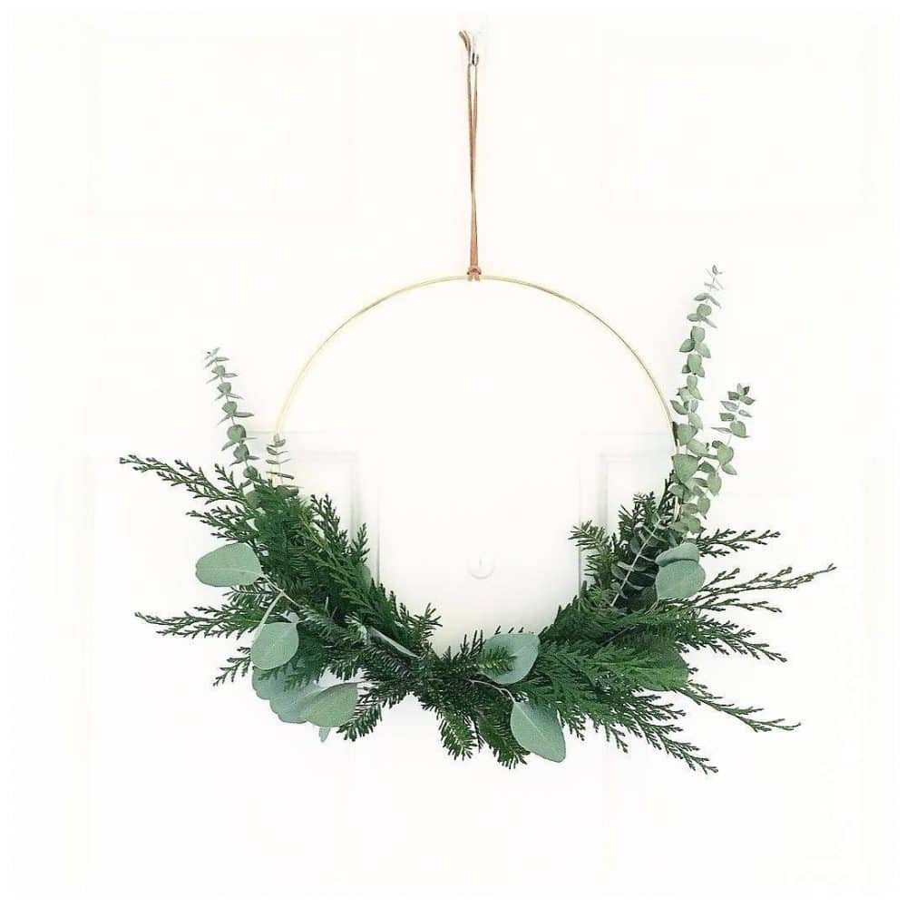 A minimalist holiday wreath with eucalyptus and evergreen branches graces a thin gold hoop, elegantly hanging from a door. The design remains simple and natural, showcasing a half-circle arrangement of lush greenery.