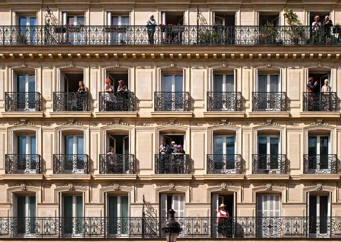 A beige building with ornate black wrought-iron balconies. People, like characters from Issue 3 of a lively urban story, stand observing the street below. The architecture features tall windows and classic Parisian design elements, stirring up the charm of vibrant city life.