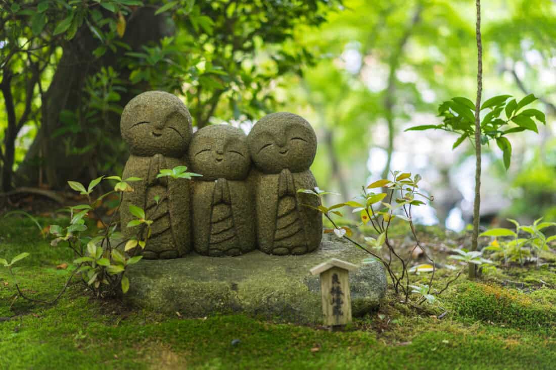 Three small, smiling stone statues with clasped hands stand together on a patch of moss and greenery in a garden. Nearby, a bamboo fountain adds melodic tranquility to the scene, enhancing the serene, natural setting provided by the surrounding plants.