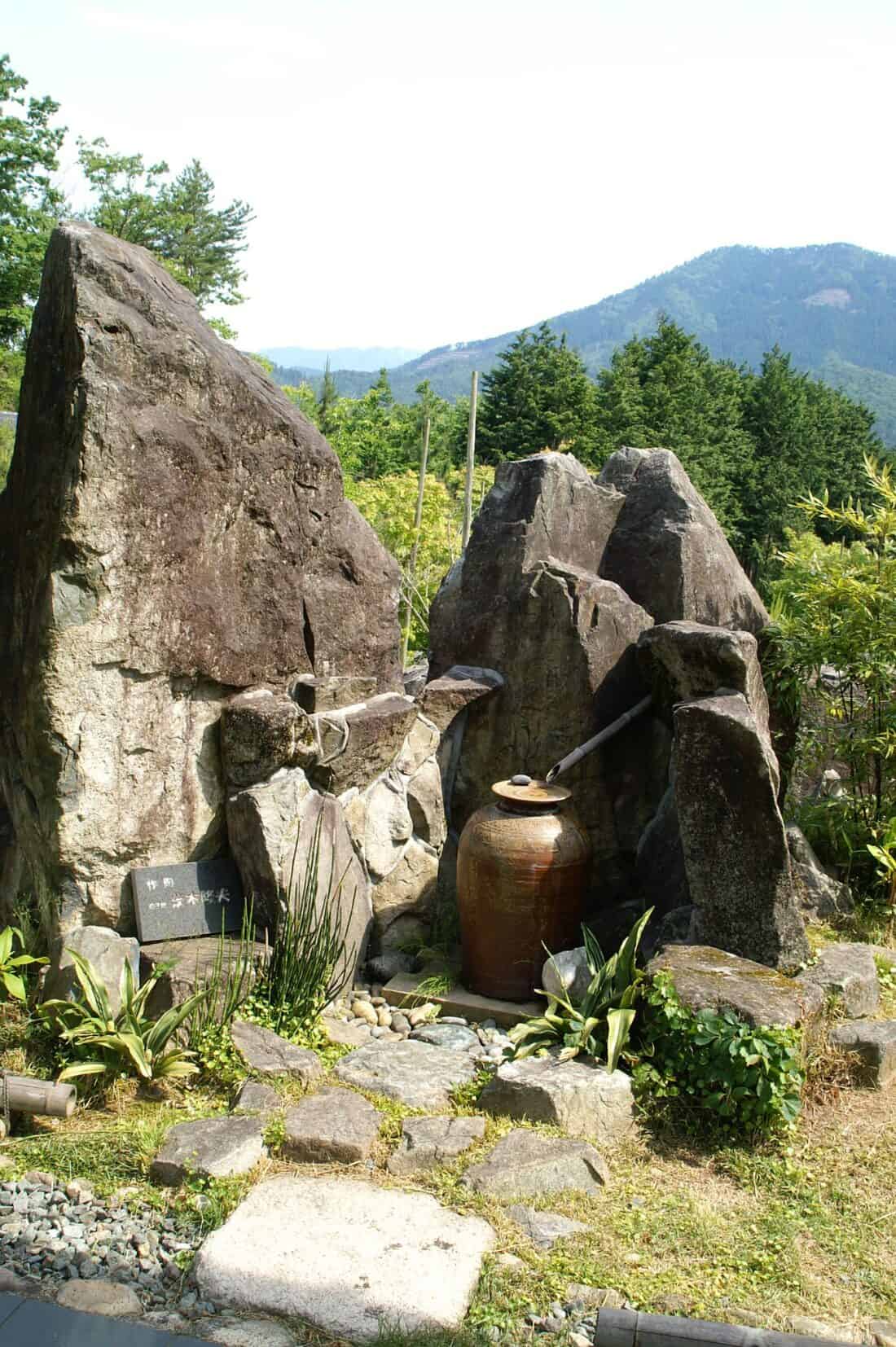 A tranquil Japanese tsukubai garden with a rustic water feature composed of large stones and a clay pot. Bamboo spouts guide water into the pot, creating a soothing cascade reminiscent of a suikinkutsu. Lush greenery and a mountain are visible in the background.