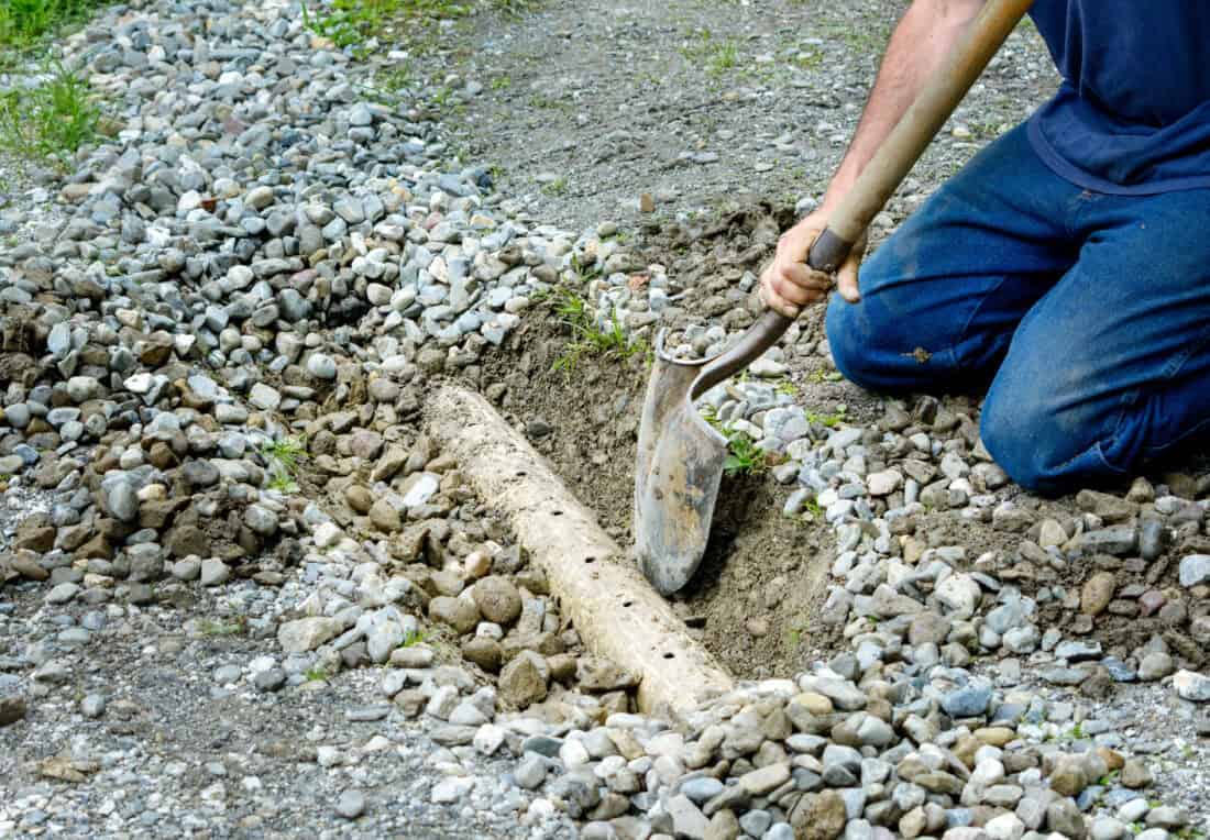 Person kneeling on gravel, using a shovel to dig a trench around a wooden log in an effort to fix a muddy backyard. The surrounding ground is rocky, and small plants are visible in the background, illustrating practical muddy backyard solutions in action.