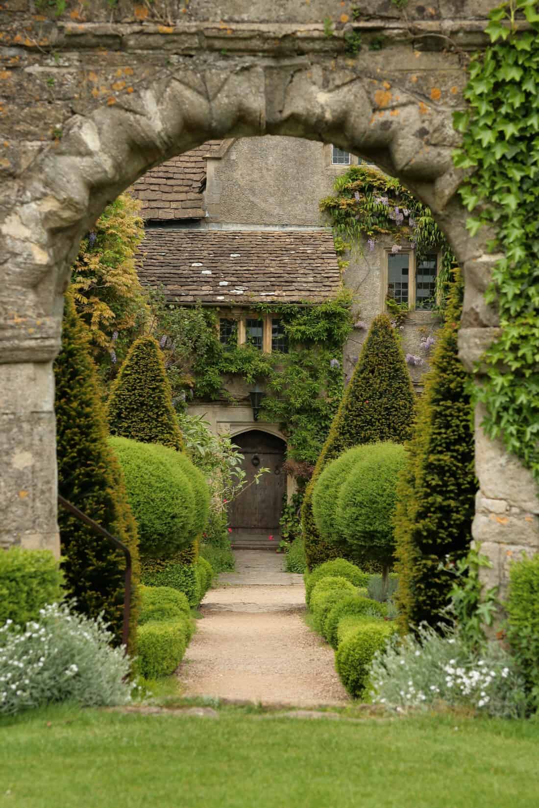 A picturesque garden pathway is framed by a stone archway, leading to an ivy-covered stone building. The path is lined with trimmed hedges and conical topiaries, creating a lush, well-maintained scene reminiscent of parterre garden elegance. The building has a rustic charm with wooden door and windows.