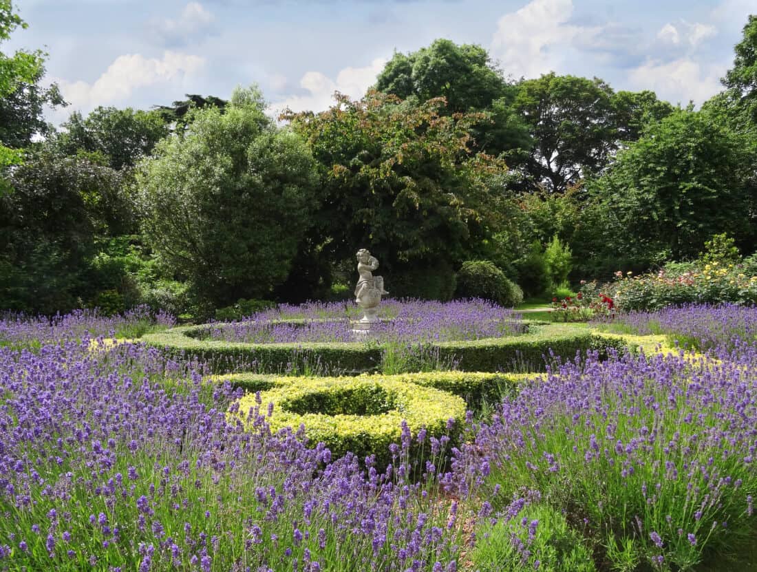 A lush garden features a central statue surrounded by manicured hedges, vibrant purple flowers, and dense green foliage. Small knot garden designs intertwine with trees and bushes, providing a dense backdrop under a partly cloudy sky. The serene scene showcases a blend of formal and natural garden elements.