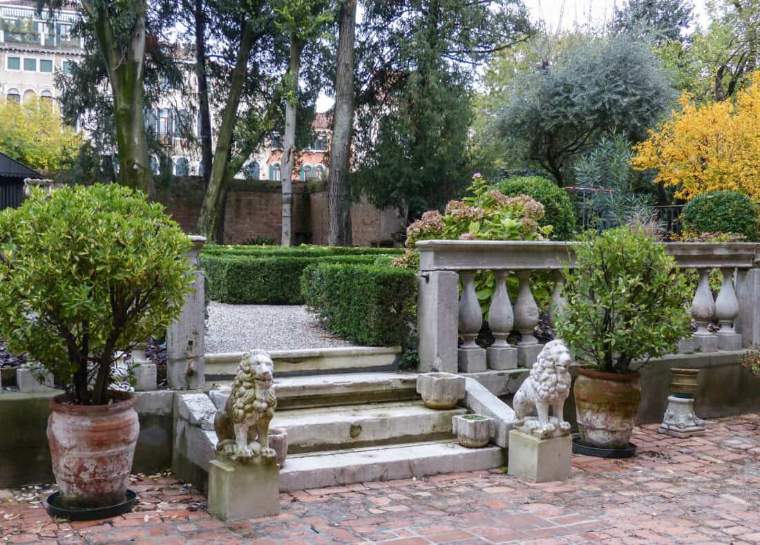 A beautifully manicured garden with stone steps leading to a gravel pathway showcases perfect scale in garden design. Two stone lion sculptures adorn the base of the steps, and potted plants flank the entrance. The scene is surrounded by lush greenery and trees, creating a serene atmosphere.