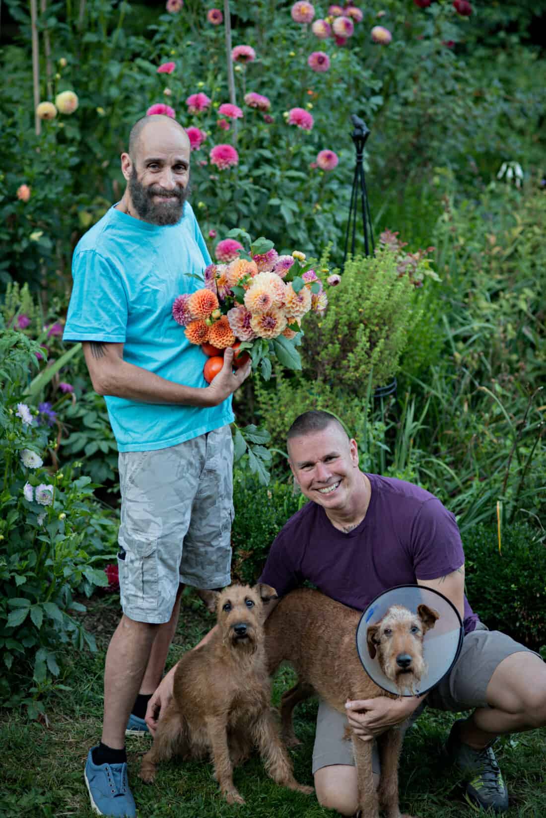 A man standing and holding a colorful bouquet stands next to another man kneeling with two dogs in a lush garden with flowers and trees in the background.