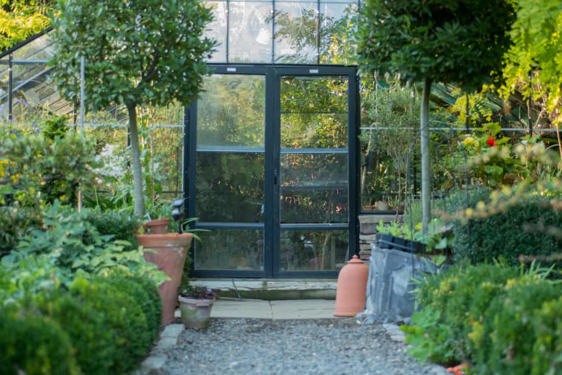 A glass greenhouse with a central black-framed door, nestled in a lush garden. potted plants line a gravel pathway leading to the entrance.