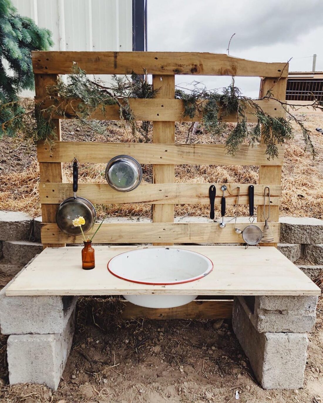 A rustic outdoor mud kitchen made from wooden pallets and concrete blocks, featuring a bowl sink, utensils, pans, and a brown bottle with a flower—perfect for creative play area ideas amid dirt and trees. A rustic outdoor mud kitchen made from wooden pallets and concrete blocks, featuring a bowl sink, utensils, pans, and a brown bottle with a flower—perfect for creative play area ideas amid dirt and trees.