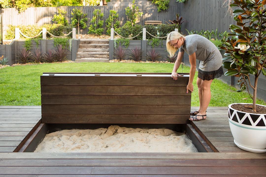 A woman lifts a wooden lid to reveal a hidden sandbox on a deck in a backyard, showcasing clever play area ideas among green grass, plants, and steps in the background. A woman lifts a wooden lid to reveal a hidden sandbox on a deck in a backyard, showcasing clever play area ideas among green grass, plants, and steps in the background.