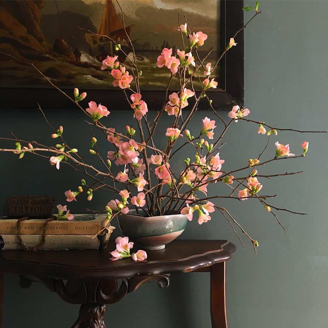 A vase of pink quince flowers sits on a table in front of a painting.