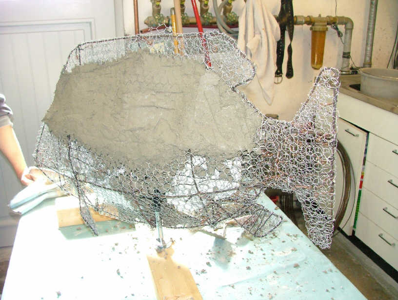 A person is working on a wire sculpture and concrete base form for a fish sculpture.