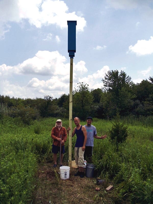 Three men standing next to a pole with bat houses in a field.