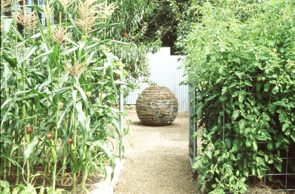 A garden pathway flanked by lush corn stalks on one side and dense greenery on the other, leading to a round stone sculpture.