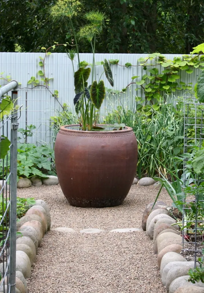 A large, round terracotta planter at the end of a garden path, surrounded by assorted green plants and a metal fence.