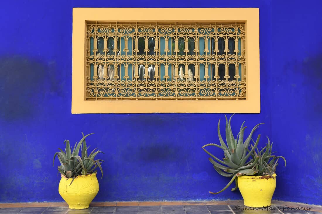 A vibrant blue wall with a decorative window framed in yellow, reminiscent of the Majorelle Garden, flanked by two large yellow pots with agave plants.