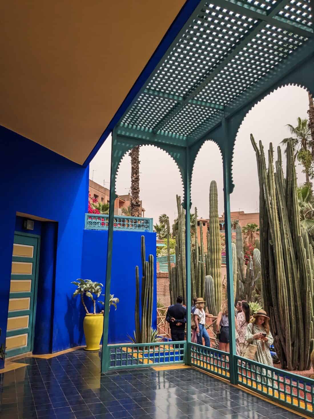 A vibrant blue courtyard with traditional moroccan architecture, featuring tall cacti and visitors exploring the space.