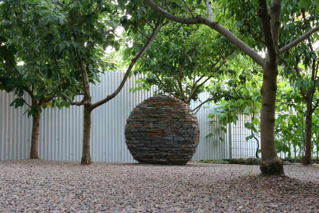 A stone sphere sculpture placed on a gravel surface with trees and a corrugated metal fence in the background.