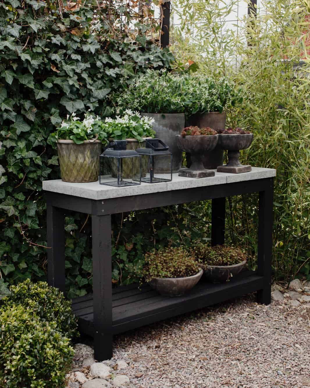 A variety of potted plants displayed on and underneath a black outdoor table, surrounded by garden foliage.