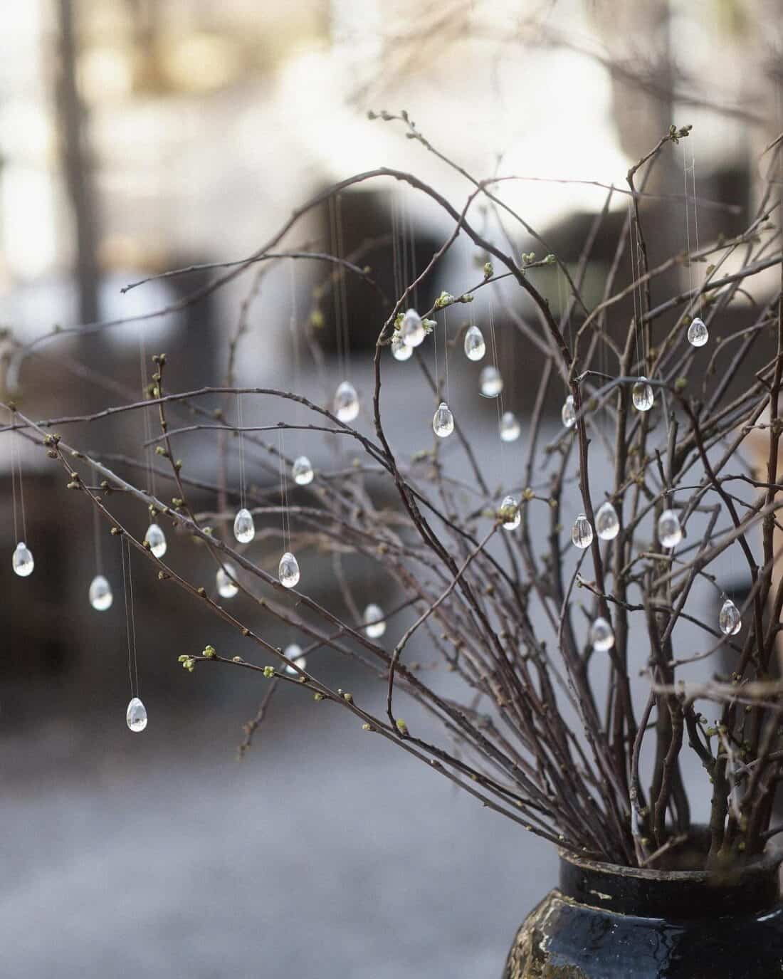 Bare twigs in a vase adorned with hanging clear glass beads, with a blurred background.