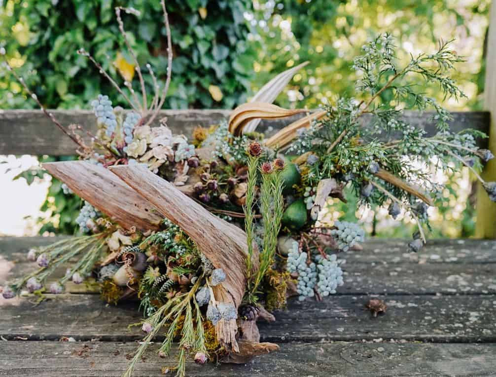 A rustic woodland garden floral arrangement with driftwood, berries, and foliage on a wooden surface.