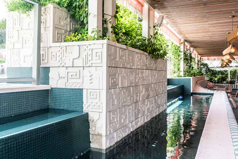 Outdoor swimming pool area featuring decorative white tiled walls, lush green plants, and a shaded seating area.