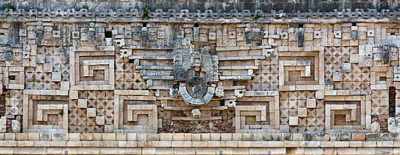 Intricate stone carvings of geometric patterns and a central ceremonial sculpture on an ancient mayan building's facade.