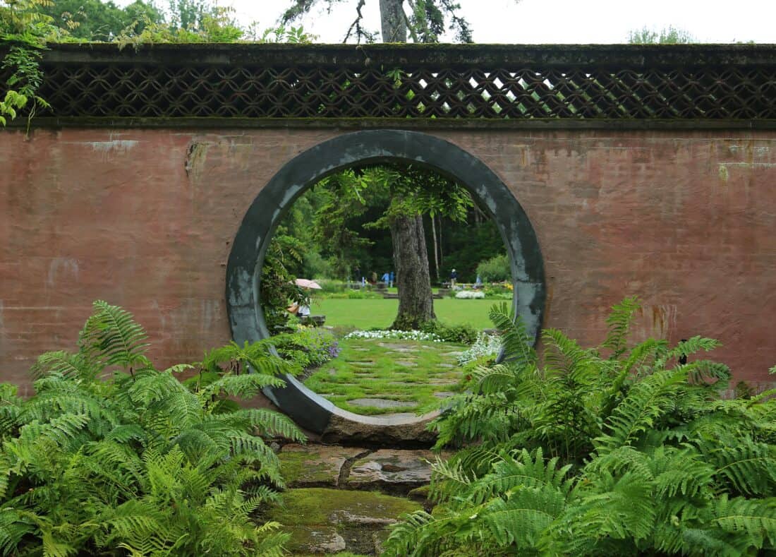 A circular stone portal in a weathered red wall, framing a view of a lush green garden with a stone path leading to a lawn dotted with people and umbrellas.