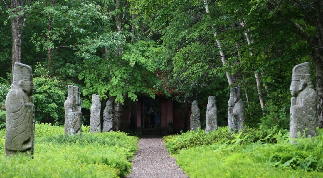 A pathway lined with ancient stone statues through a lush forest leading to a distant building.