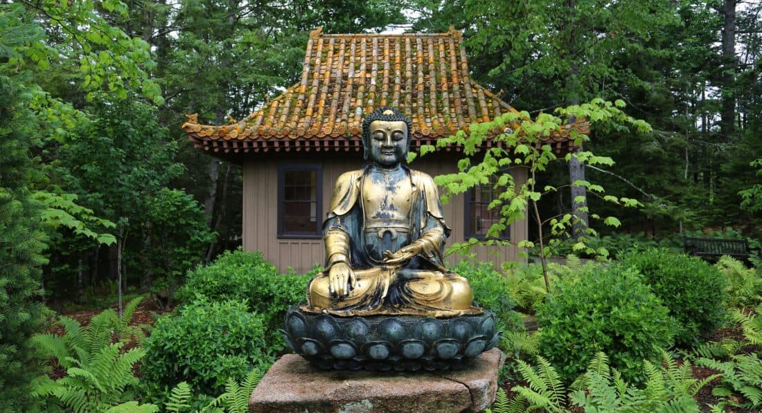 Bronze buddha statue seated in lotus position under a small, traditional asian-style pavilion amidst lush greenery.