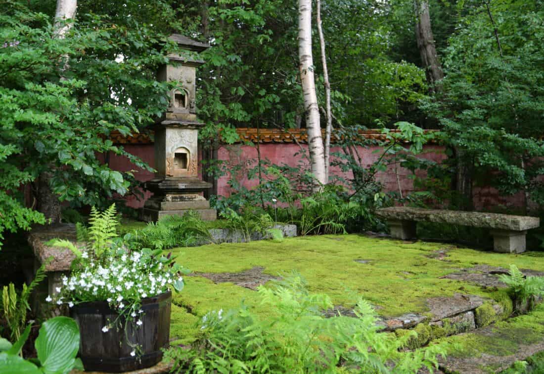 Old moss-covered stone pagoda in a lush green garden with a wooden bench, surrounded by trees and a potted plant.