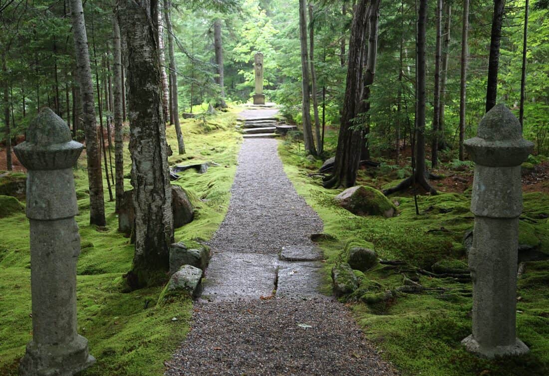 A gravel pathway flanked by moss-covered stone pillars, leading through a lush green forest to a statue in the distance.