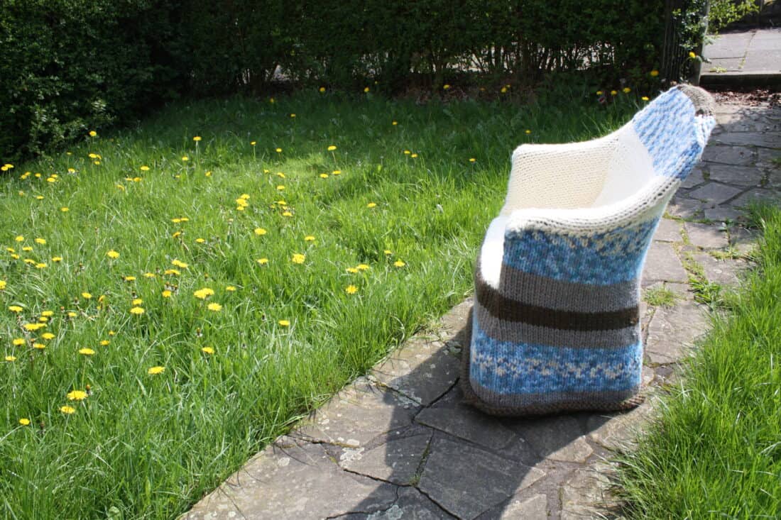 A knitted blanket draped over a garden chair on a lawn with dandelions.