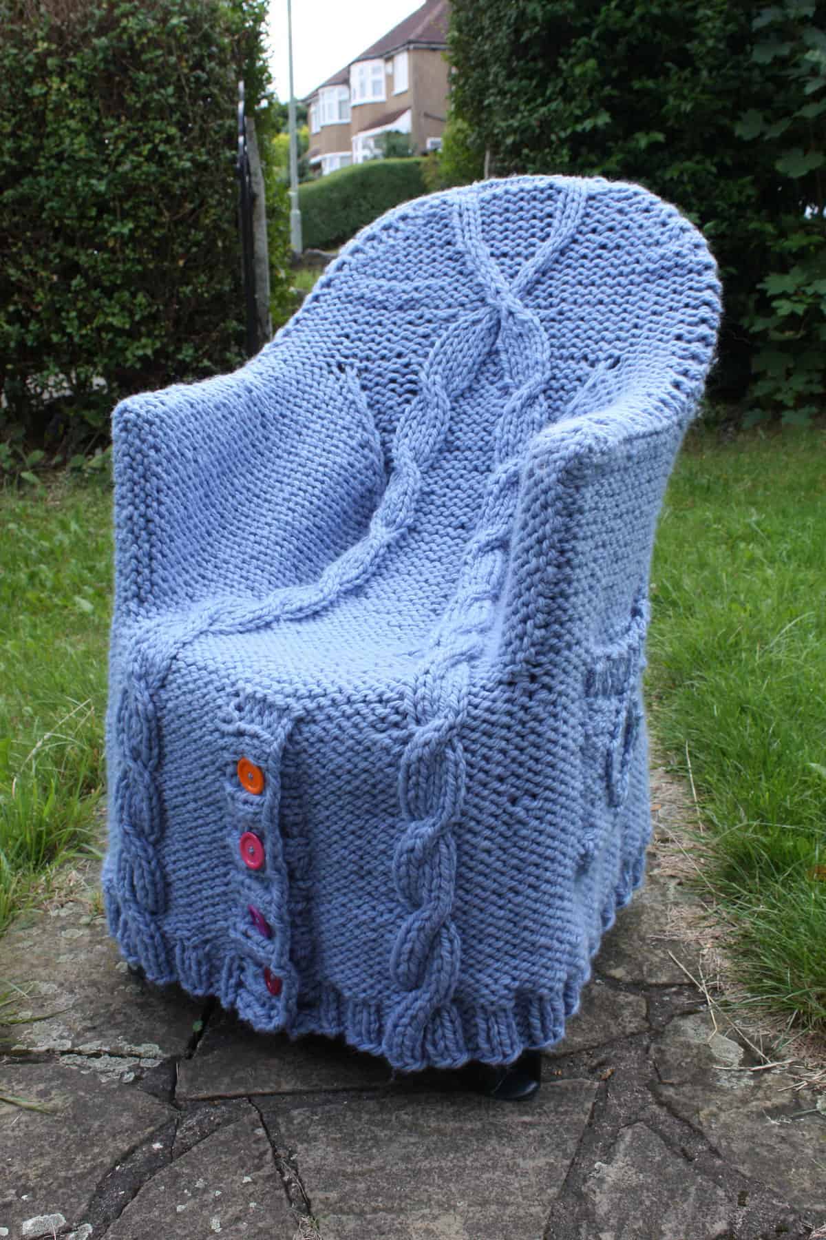 A knitted chair cover with cable patterns and buttons displayed outdoors.