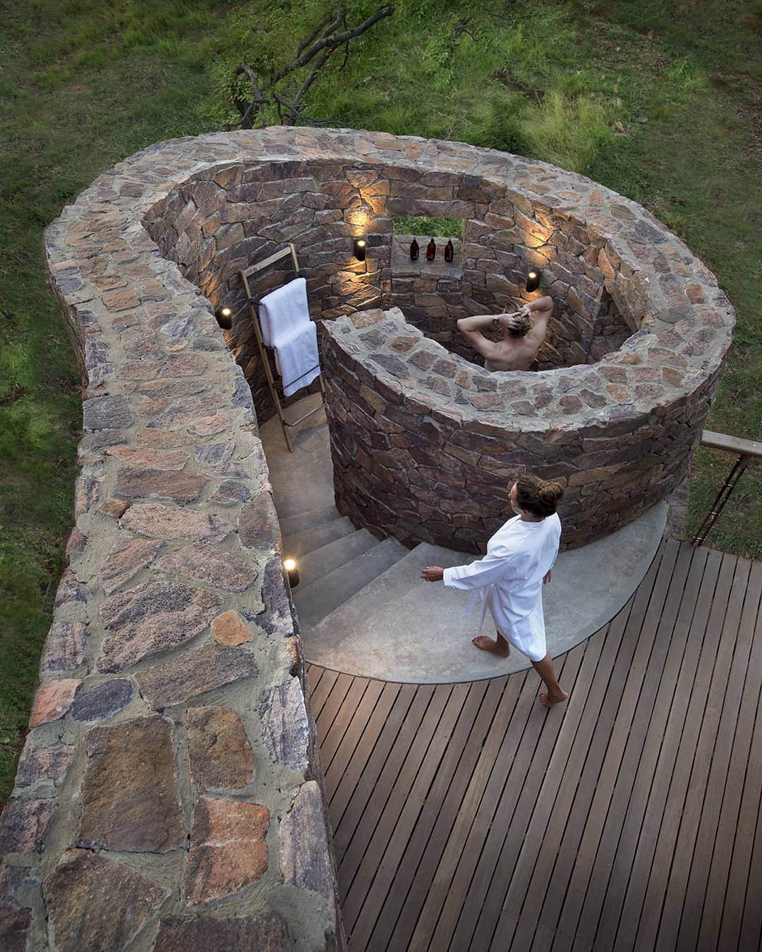 A woman in a white robe walks towards an elevated stone-built circular spa bath surrounded by greenery, where another woman relaxes; an exhaustive roundup of outdoor shower ideas enhances the serene ambiance.