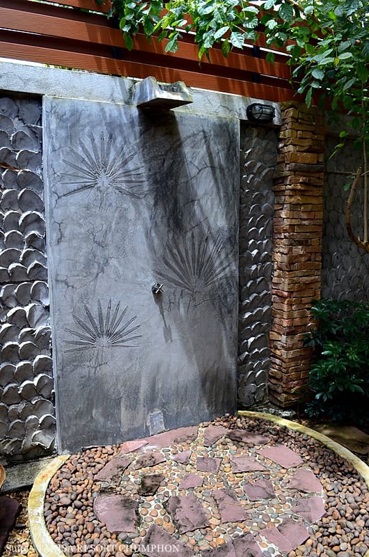 A decorative outdoor shower with a unique textured wall adorned with leaf patterns, surrounded by lush green plants and stone tiles, perfect for a cool-off.