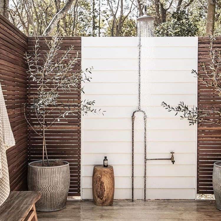Outdoor shower with a white panel backdrop and wooden privacy fence, designed for those looking to cool off, featuring a suspended shower head surrounded by potted plants and a rustic wooden bench.