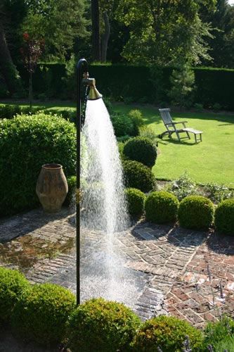 Outdoor shower ideas with a strong stream of water cascading down, situated on a stone patio surrounded by lush greenery and a manicured lawn with a solitary sun chair in the background.