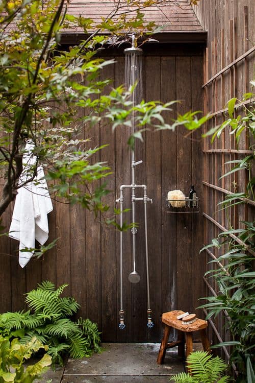 An outdoor shower surrounded by lush greenery with a wooden stool and hanging white towel provides an ideal spot to cool-off. The shower, with its metallic finish, is attached to a wooden wall, enhancing