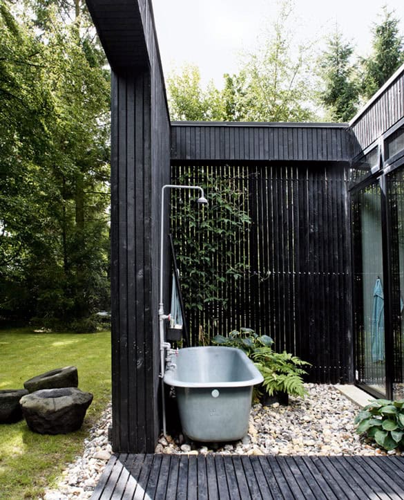 Outdoor shower setup featuring a large, freestanding metal bathtub against a modern black slatted wooden wall, surrounded by lush greenery and pebble ground, ideal for a cool-off.