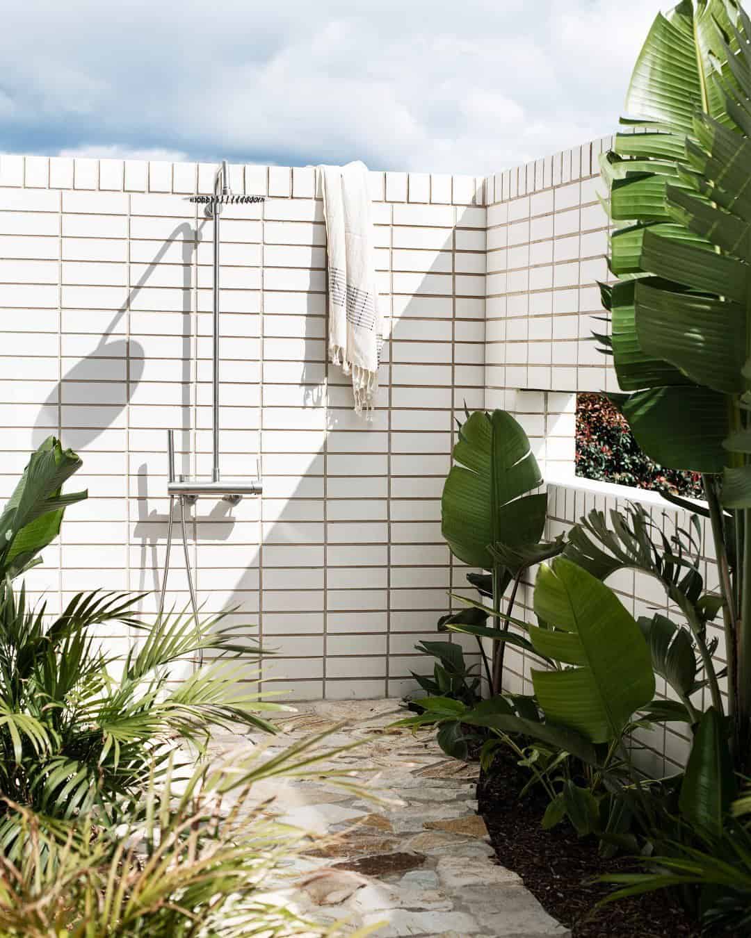 Outdoor shower area surrounded by white tile walls embellished with green tropical plants, offering a cool-off spot with a towel hanging on a steel hook, under a partly cloudy sky.